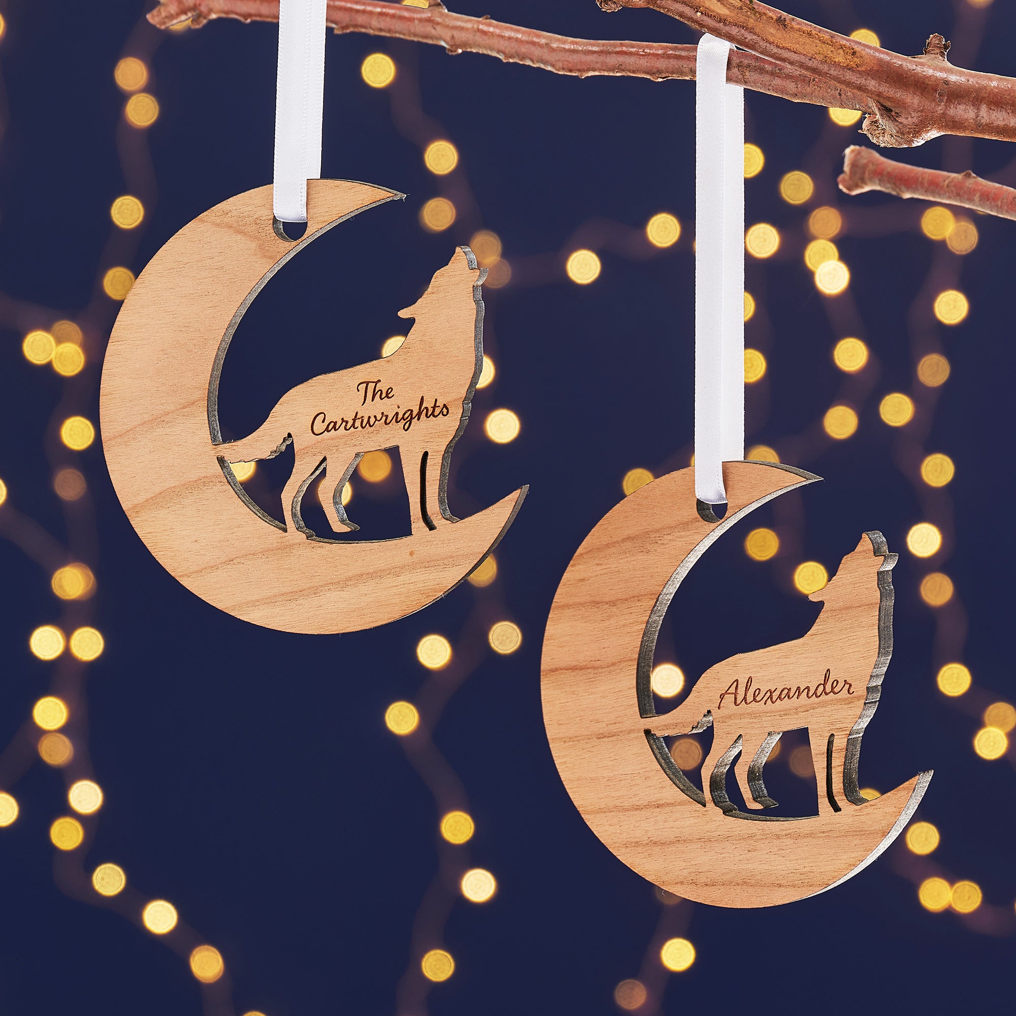 Two wooden Christmas ornaments, one a crescent moon and the other a circle, hang from a branch against a dark blue background with bokeh lights. Each ornament features a cutout silhouette of a wolf howling at the moon and is engraved with a family name, "The Cartwrights" and "The Smiths."