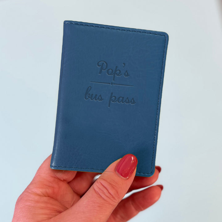 A womans hand holds a blue custom ID holder/bus pass leather holder. The front of the slim style wallet has been personalised with the words Pop's bus pass.