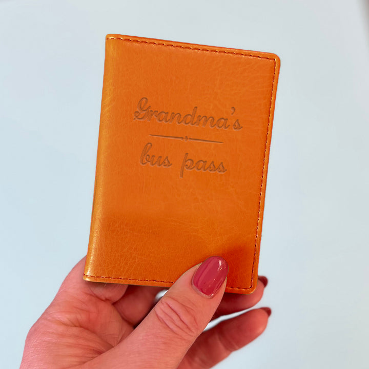 A womans hand holds an orange custom ID holder/bus pass leather holder. The front of the slim style wallet has been personalised with the words Grandma's bus pass.