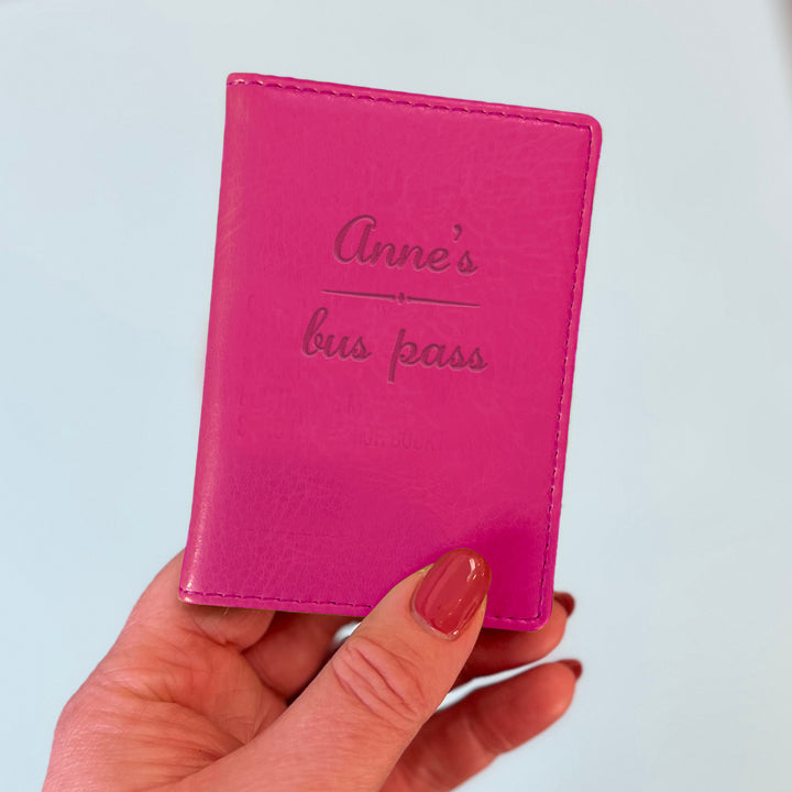 A womans hand holds a pink custom ID holder/bus pass leather holder. The front of the slim style wallet has been personalised with the words Anne's bus pass.