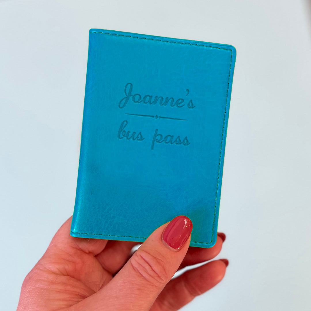A womans hand holds a turquoise custom ID holder/bus pass leather holder. The front of the slim style wallet has been personalised with the words Joanne's bus pass.