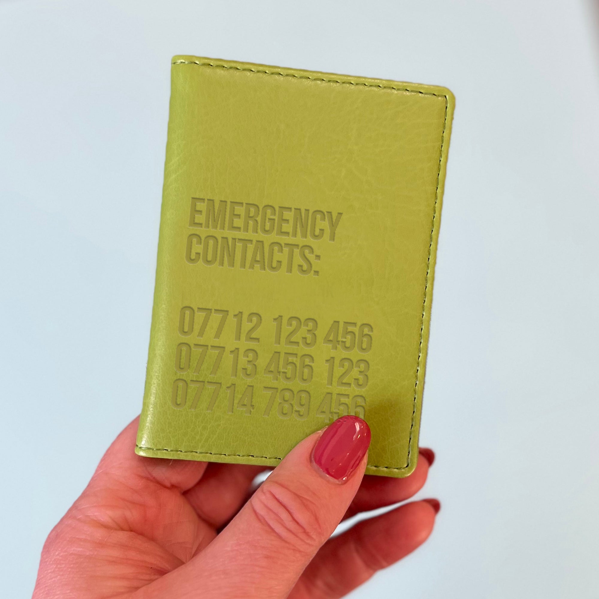 A womans hand holds a green custom ID holder/bus pass leather holder. The front of the slim style wallet has been personalised with the words Emergency contacts and then three mobile phone numbers.