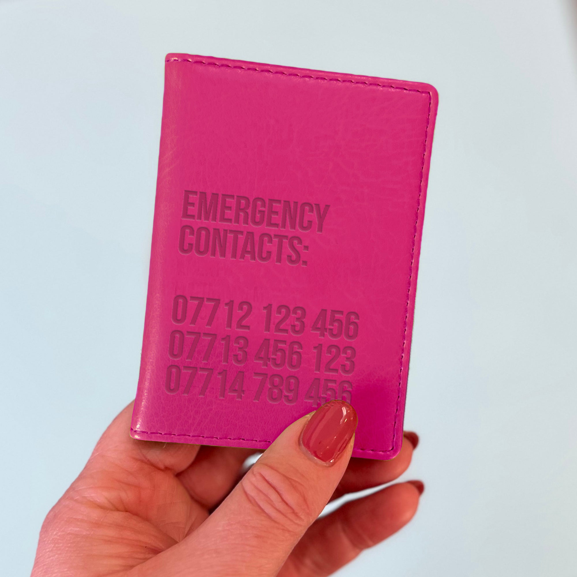 A womans hand holds a pink custom ID holder/bus pass leather holder. The front of the slim style wallet has been personalised with the words Emergency contacts and then three mobile phone numbers.