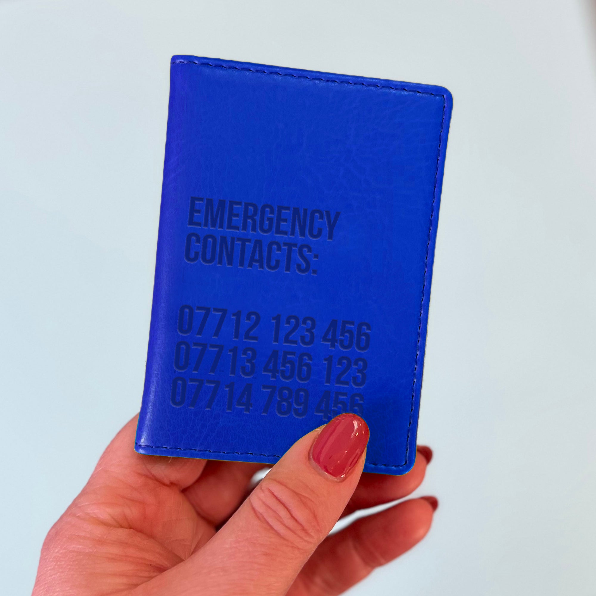 A womans hand holds a royal blue custom ID holder/bus pass leather holder. The front of the slim style wallet has been personalised with the words Emergency contacts and then three mobile phone numbers.