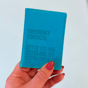 A womans hand holds a turquoise custom ID holder/bus pass leather holder. The front of the slim style wallet has been personalised with the words Emergency contacts and then three mobile phone numbers.