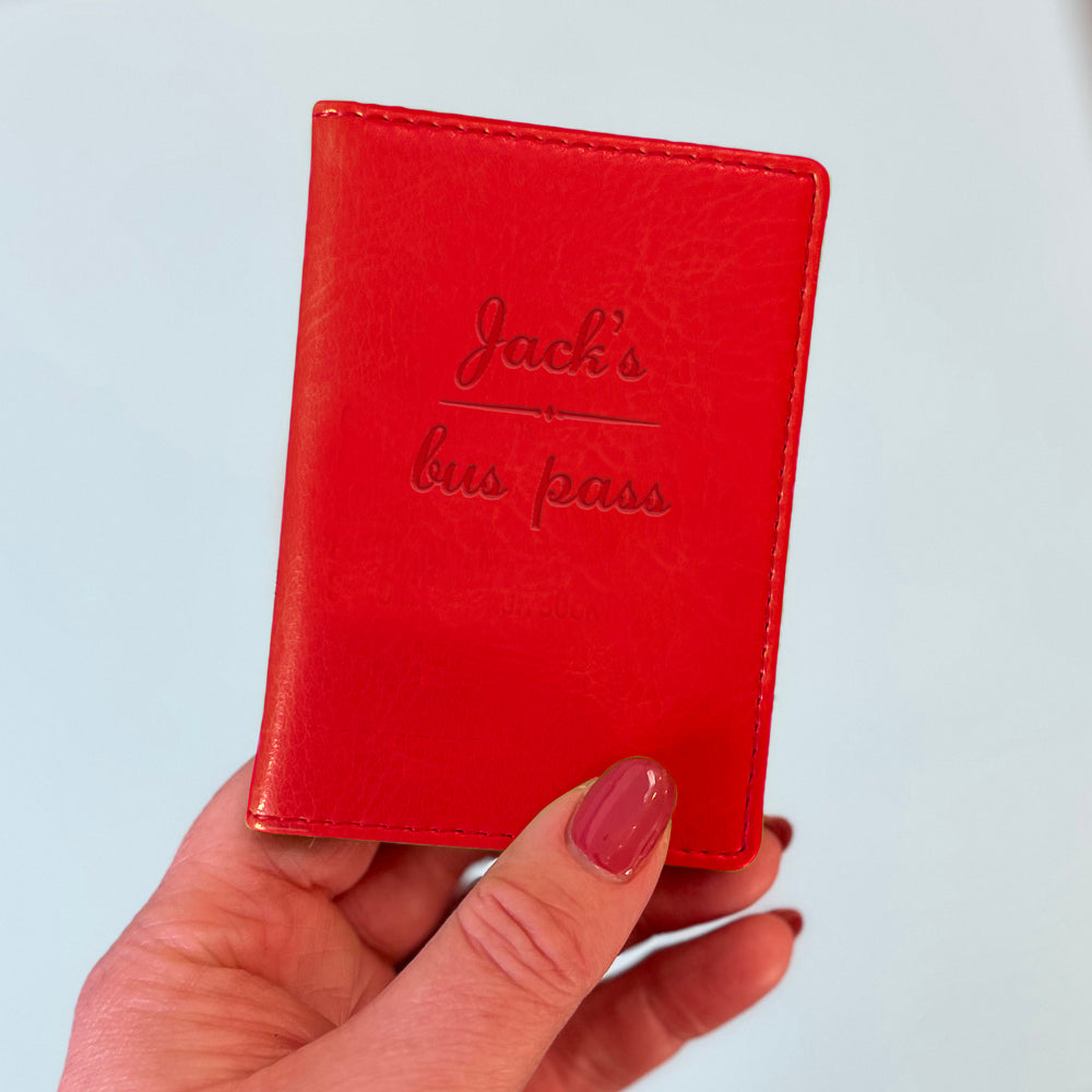 A womans hand holds a red custom ID holder/bus pass leather holder. The front of the slim style wallet has been personalised with the words Jack's bus pass.