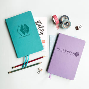 A top down image of two vegan leather notebooks, one turquoise and one lilac. They are resting on a pale aqua table. The front of each notebook is personalised with an engraving of a business logo.  Surrounding the book are various stationary items.