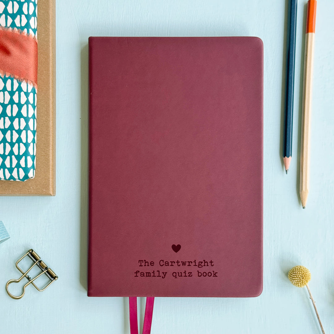 A top down image of a Burgundy hardcover vegan leather notebook resting on a pale aqua table. The bottom of the book is engraved with a personalised message, The Cartwright family quiz book and a solid heart symbol is engraved above the text. Surrounding the book are various stationary.