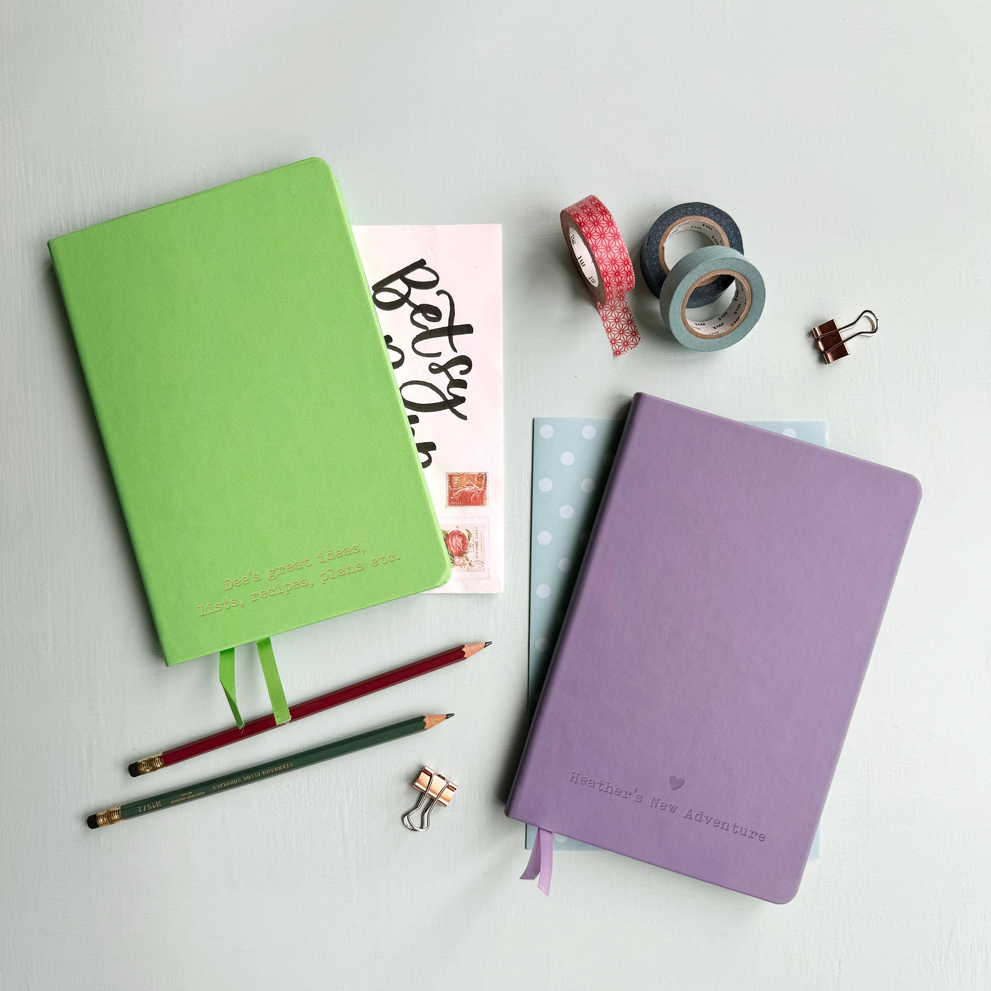 A top down image of two vegan leather notebooks, one green and one lilac. They are resting on a pale aqua table. The front of the green notebook is engraved with the personalised text “ Dee’s great ideas, lists, recipes, plans etc”. The lilac notebook is engraved with a heart and the bespoke text reads  “Heather’s New Adventure”. Surrounding the book are various stationary items.
