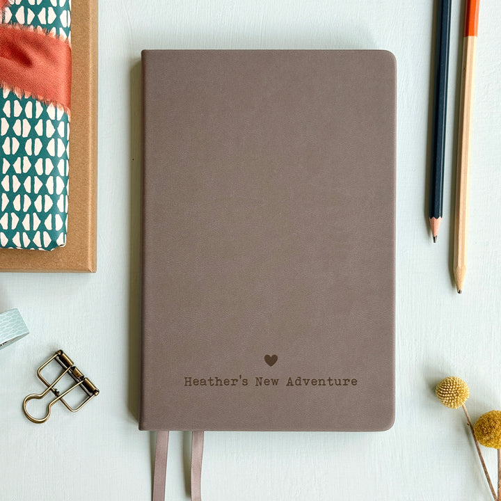 A top down image of a Grey hardcover vegan leather notebook resting on a pale aqua table. The bottom of the book is engraved with a personalised message, Heather's New Adventure and a solid heart symbol is engraved above the text. Surrounding the book are various stationary.