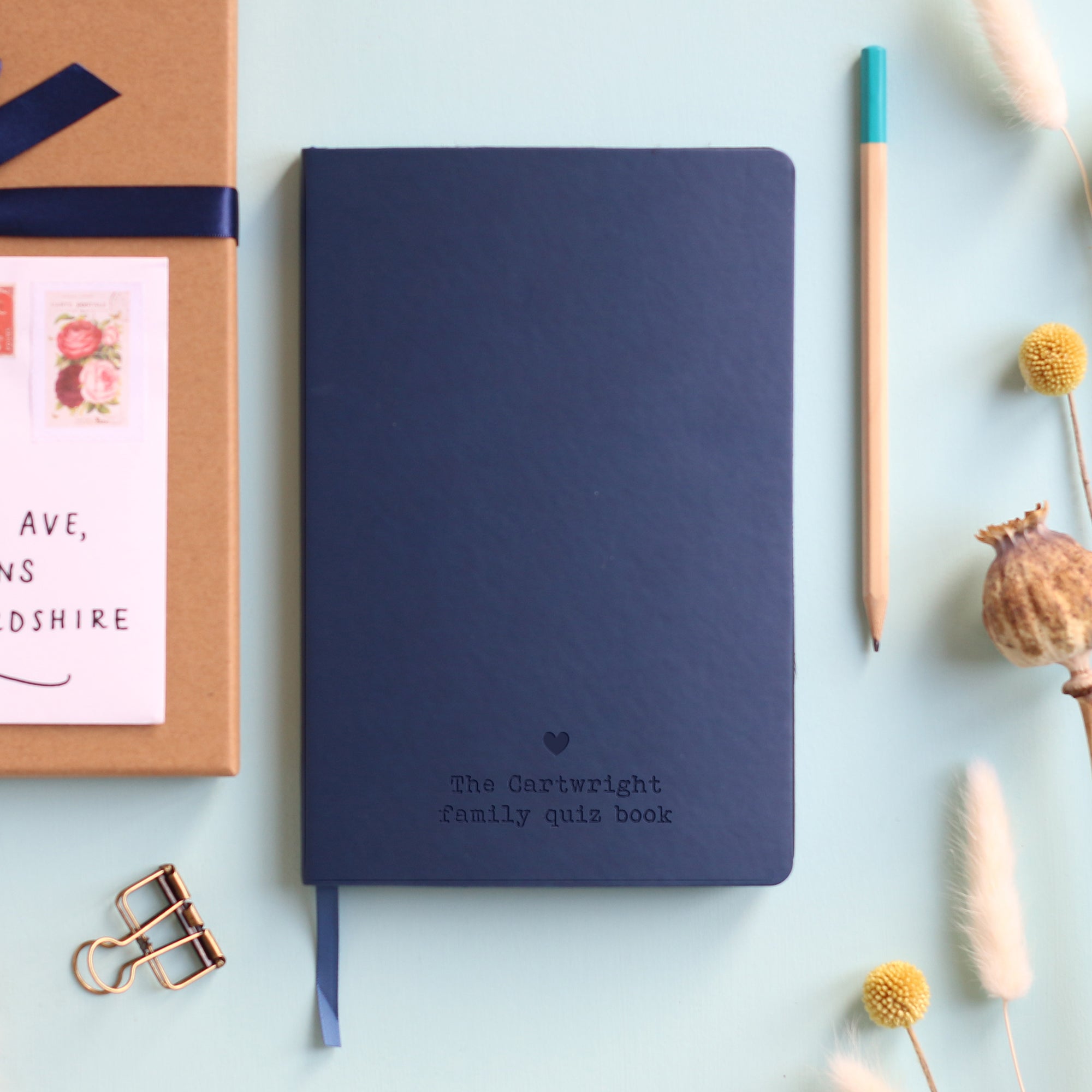 A top down image of a navy blue hardcover vegan leather notebook resting on a pale aqua table. The bottom of the book is engraved with a personalised message, "The Cartwright family quiz book" and a solid heart symbol  is engraved above the text. Surrounding the book are various stationary items and dried flowers.