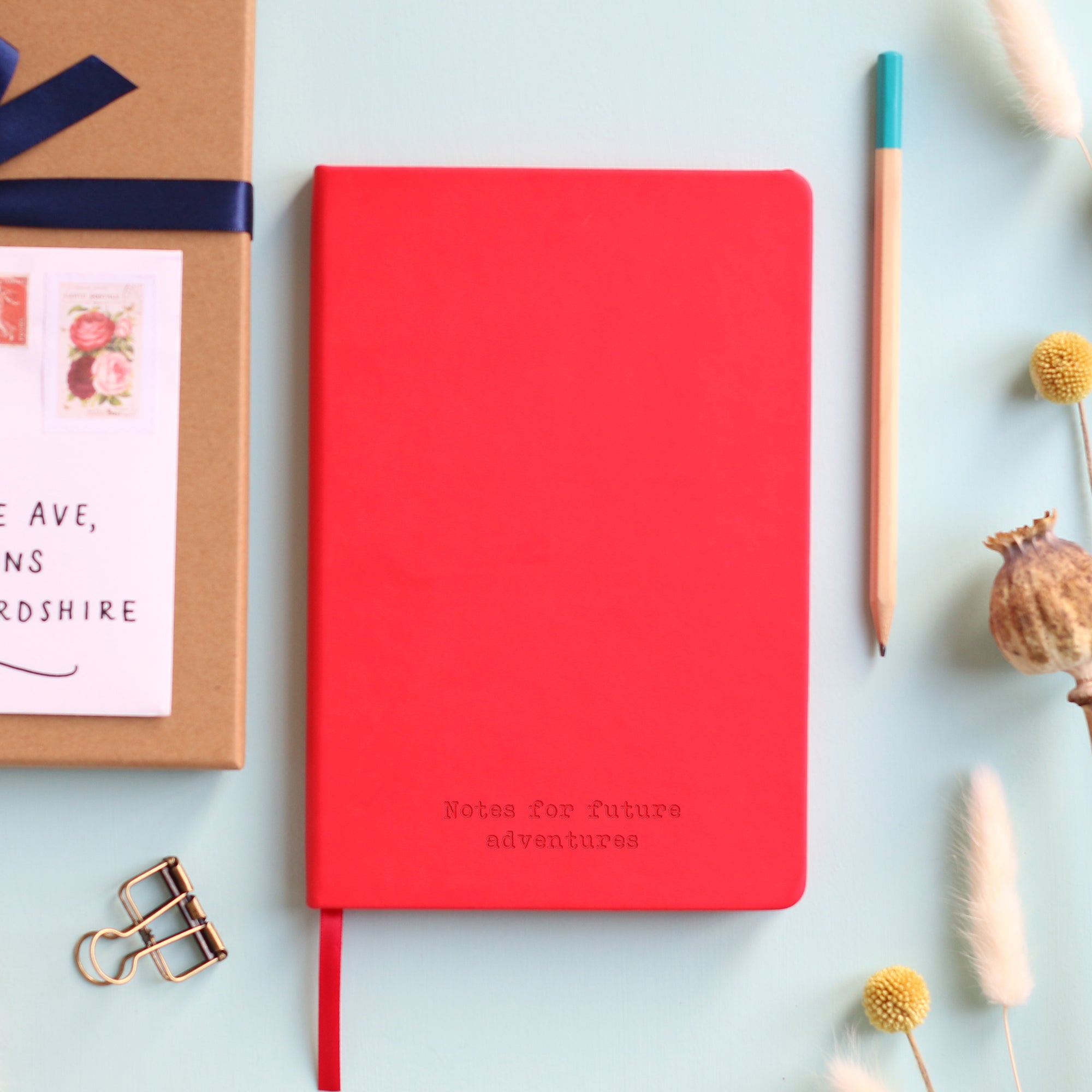A top down image of a red hardcover vegan leather notebook resting on a pale aqua table. The bottom of the book is engraved with a personalised message, "Notes for future adventures". Surrounding the book are various stationary items and dried flowers.