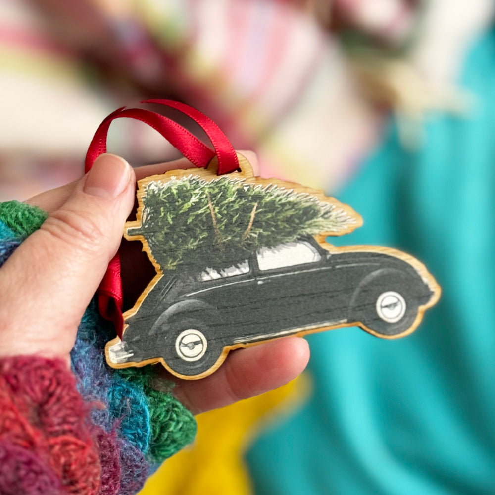 A hand holding a wooden Christmas ornament shaped like a vintage black Volkswagen Beetle with a Christmas tree strapped to the roof.