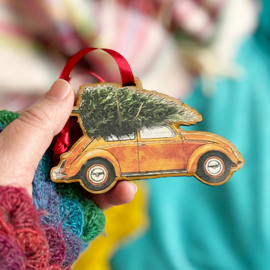 A hand holding a wooden Christmas ornament shaped like a vintage yellow Volkswagen Beetle with a Christmas tree strapped to the roof.