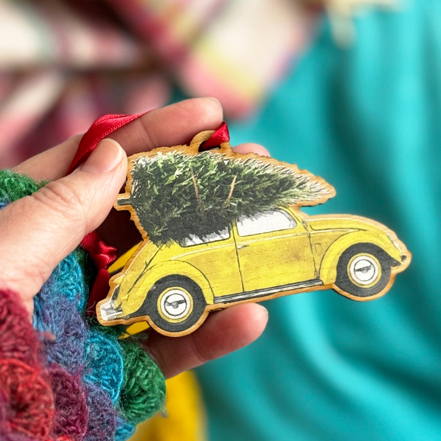 A hand holding a wooden Christmas ornament shaped like a yellow Volkswagen Beetle with a Christmas tree strapped to the roof.