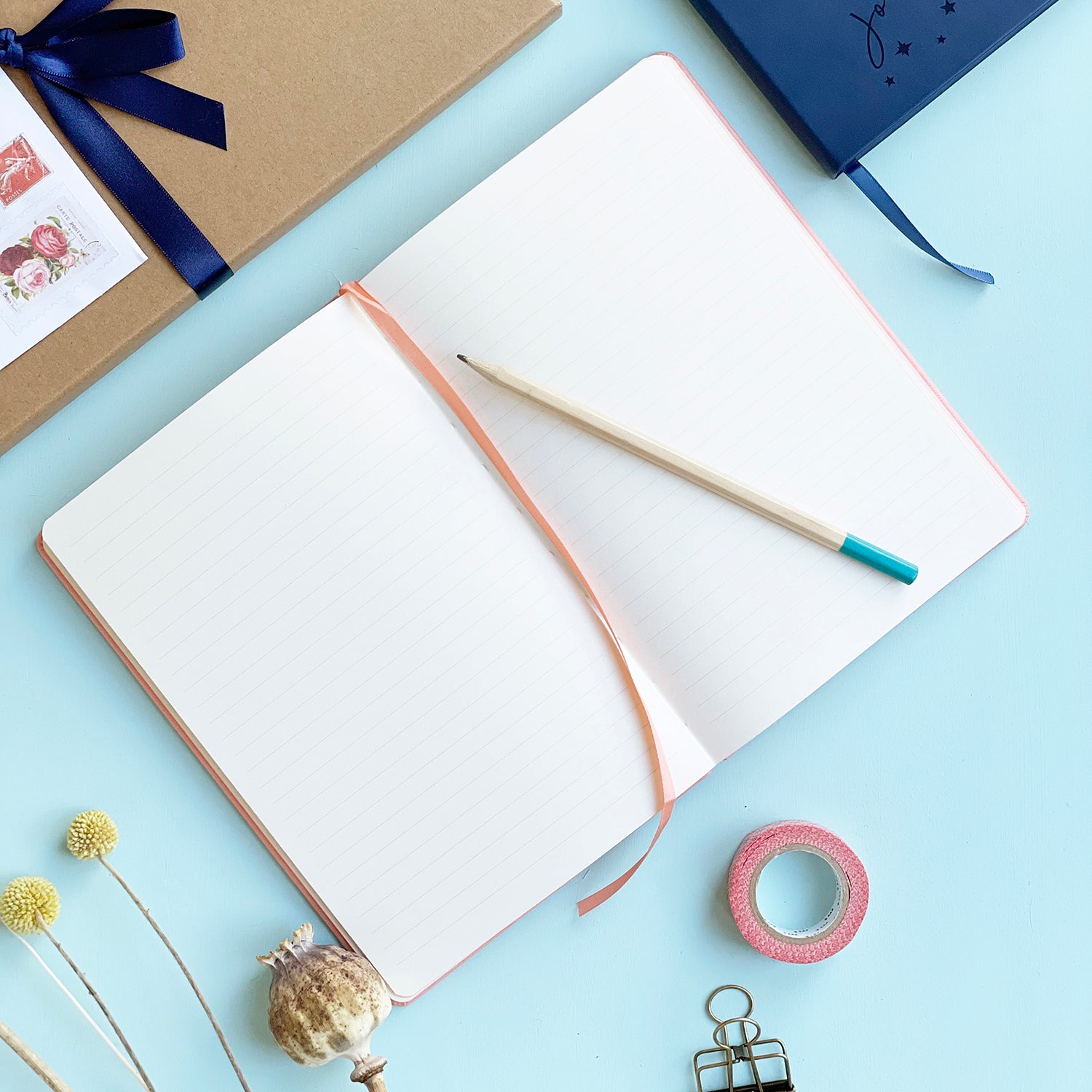  Personalised journal notebook lying flat open on pale aqua table to show lined pages and a matching colour ribbon place marker. A pencil is resting on the top of the notebook. Washi tape, clips and other stationery items are scattered around it. 