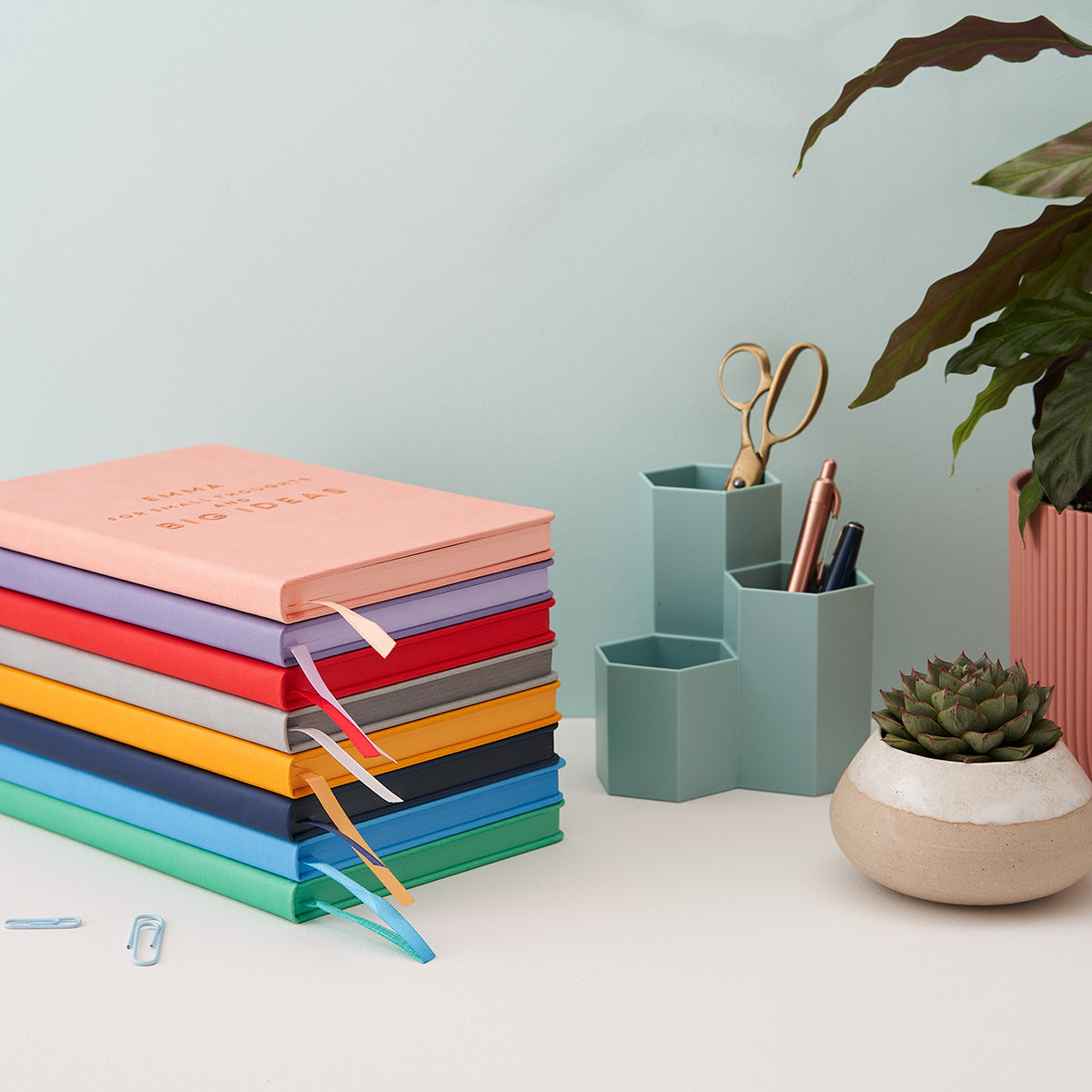 Colourful stack of leather notebooks showing the paper edges, and ribbon place markers have same colour as the cover of the book. Stack is sitting on a white desk in front of a pale aqua wall. Next to the stack is a pen pot with pens and scissors, and some potted house plants