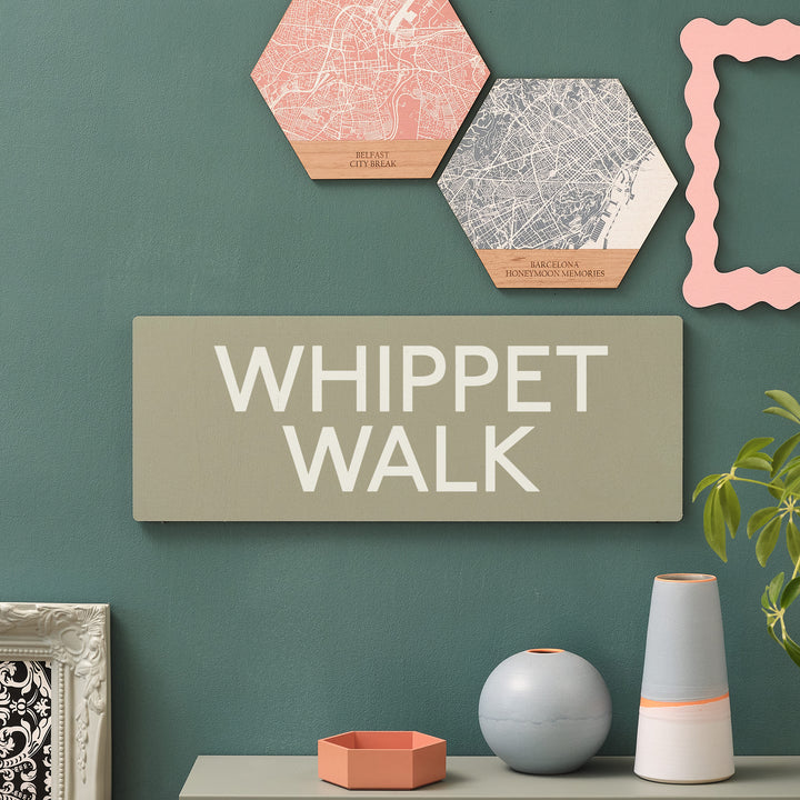 Personalised dog walker gift for pet lovers wooden wall art. White bold graphic text stating 'Whippet Walk' on a sage green wooden rectangle with rounded corners for a maximalist modern art vibe. On a forest green wall with hexagonal wooden map wall art print signs above and ceramic vases below.