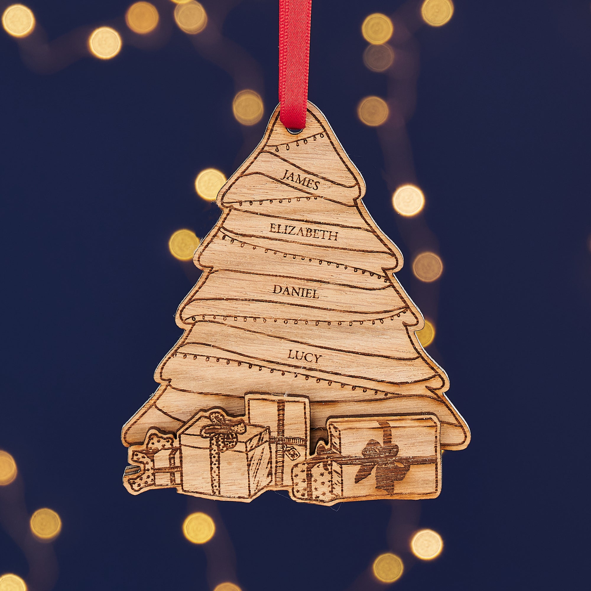 Two personalized wooden Christmas tree ornaments with engraved names, hanging from red ribbons against a background of warm fairy lights.