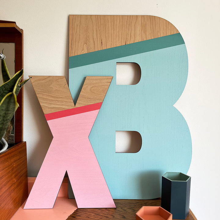 A vibrant and playful display featuring large wooden letters "X" and "B" painted in a striking color palette of pink, teal, and natural wood tones, displayed on a shelf with decorative objects.