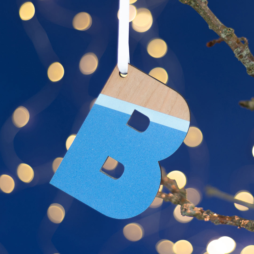 A cheerful Christmas ornament featuring the letter "B" cut from wood and painted in a vibrant shade of blue with a natural wood accent. The ornament is hanging from a branch against a backdrop of twinkling lights, adding a festive touch.
