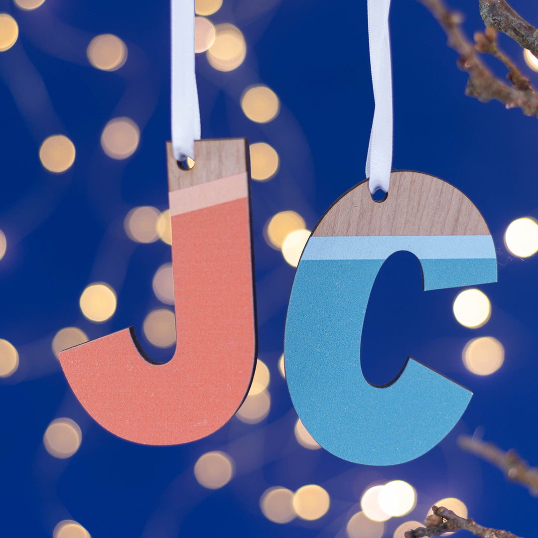A pair of cheerful Christmas ornaments featuring the letters "J" and "C" cut from wood and painted in vibrant colors. The ornaments are hanging from a branch against a backdrop of twinkling lights, adding a festive touch.