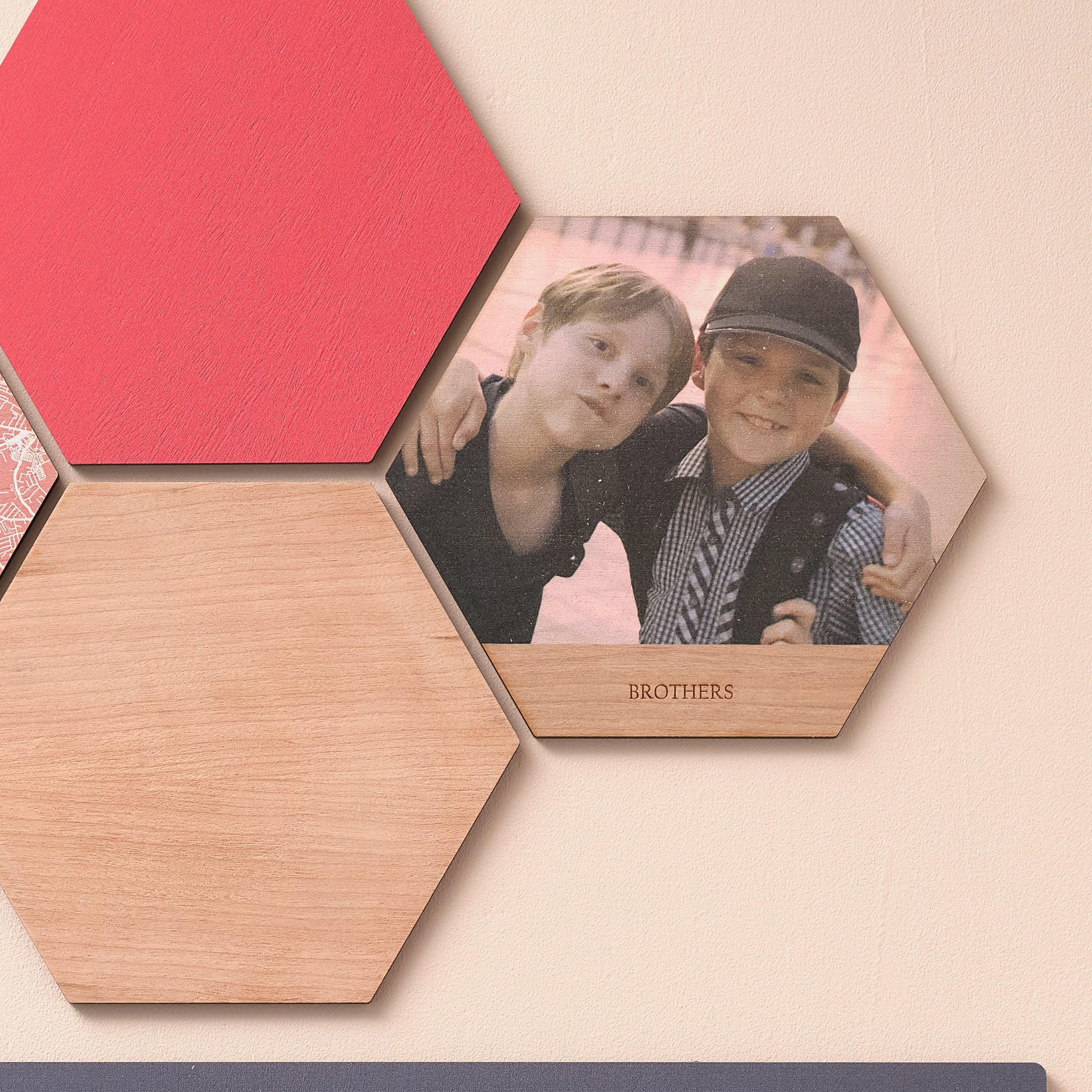 A close up of three wooden hexagonal wall art tiles. One tile shows two boys with a line of text engraved that says "brothers". One tile is solid red, one tile is plain wood.