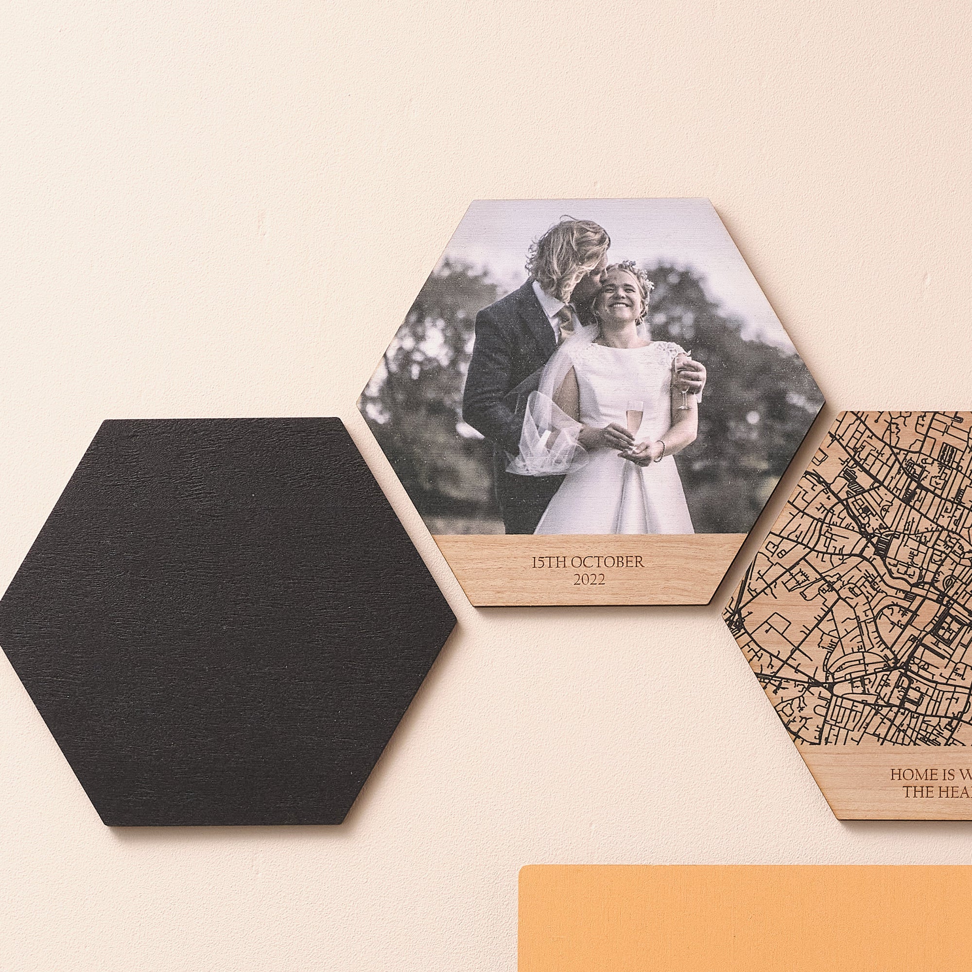 Three wooden photo tiles showing a solid black colour, a wedding photo with personalised tex, and a black line map. Hung on a pink wall.