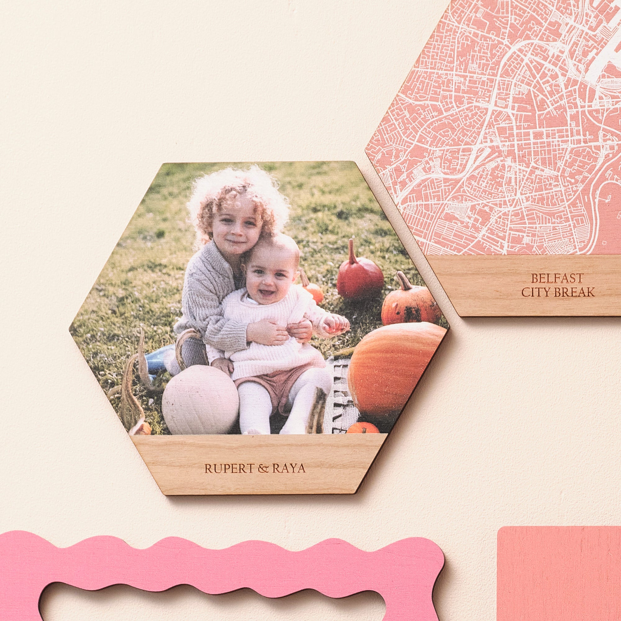 Two wooden hexagonal wall art tiles on a pink wall. One shows a photograph of two children in a pumpkin patch, one shows a white map on a pink background. Both have lines of engraved text beneath.