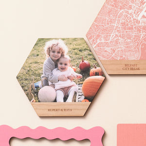 Two wooden hexagonal wall art tiles on a pink wall. One shows a photograph of two children in a pumpkin patch, one shows a white map on a pink background. Both have lines of engraved text beneath.