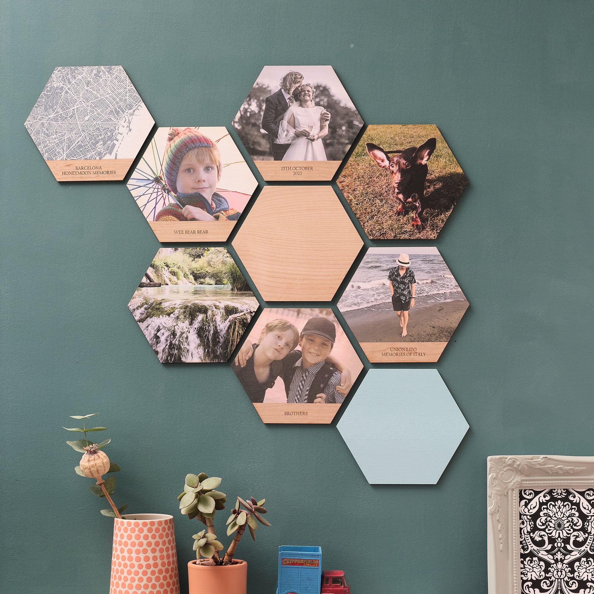 A collage of hexagonal wooden tiles featuring photos, maps, and solid colours. Some tiles also have personalised engraved text.  Mounted on a green wall.