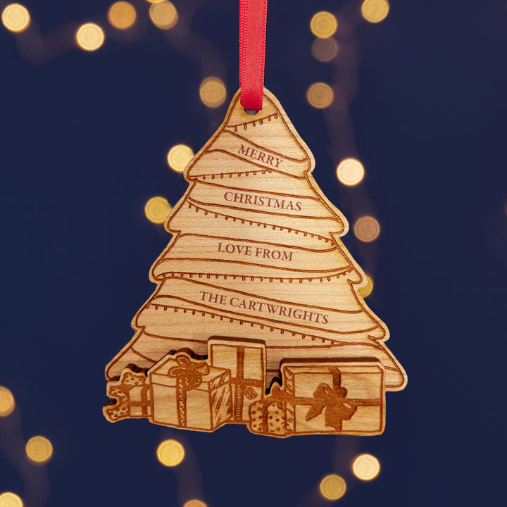 A personalized wooden Christmas tree ornaments with engraved names, hanging from red ribbons against a background of warm fairy lights.