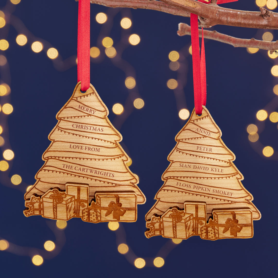 Two personalized wooden Christmas tree ornaments with engraved names, hanging from red ribbons against a background of warm fairy lights.