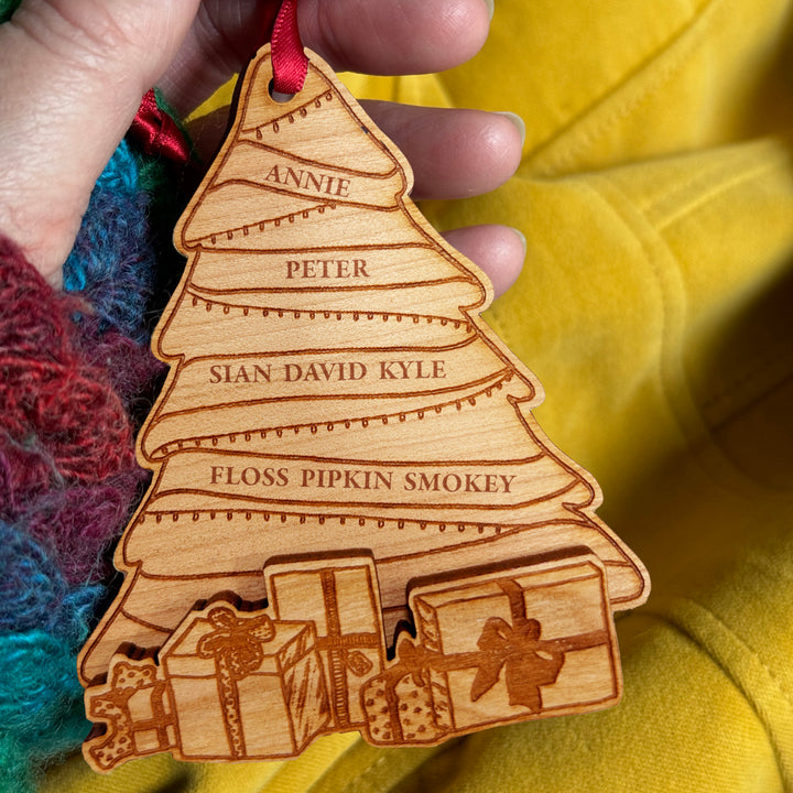 A personalized wooden Christmas tree ornament with engraved names, held in a hand with a red ribbon loop, against a yellow fabric background.