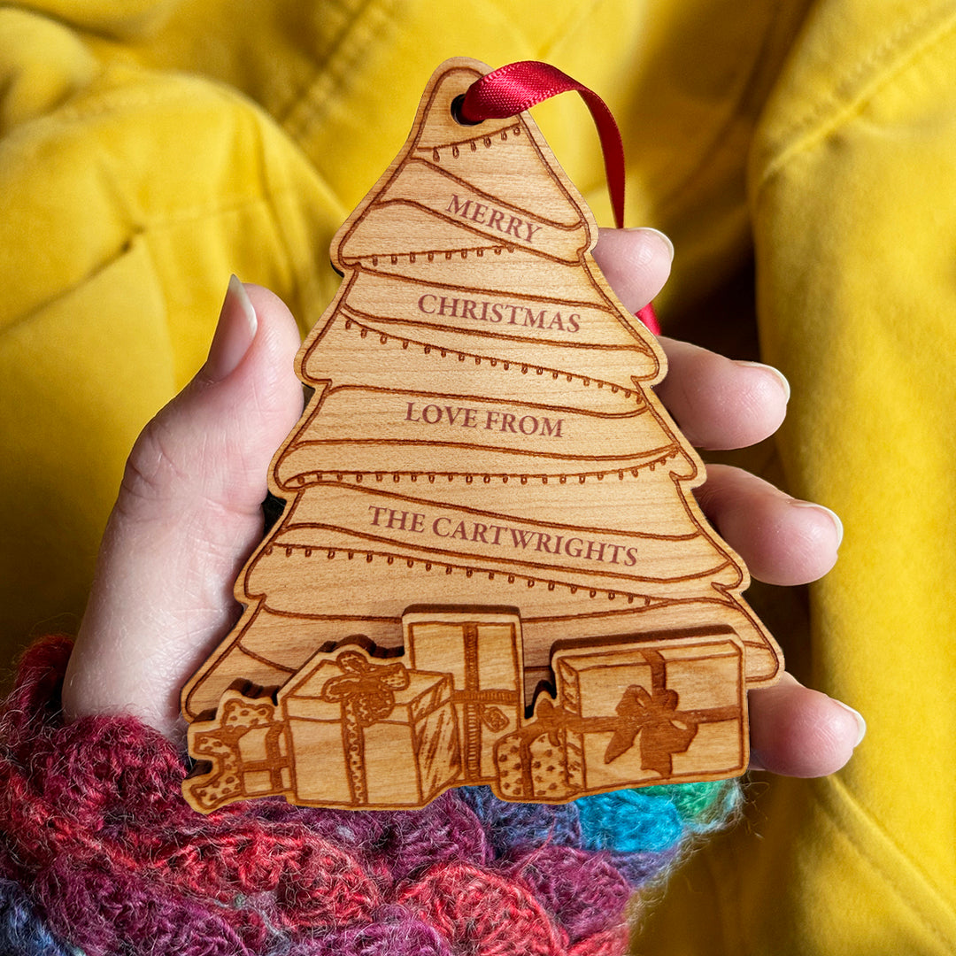A personalized wooden Christmas tree ornament with engraved names, held in a hand with a red ribbon loop, against a yellow fabric background.