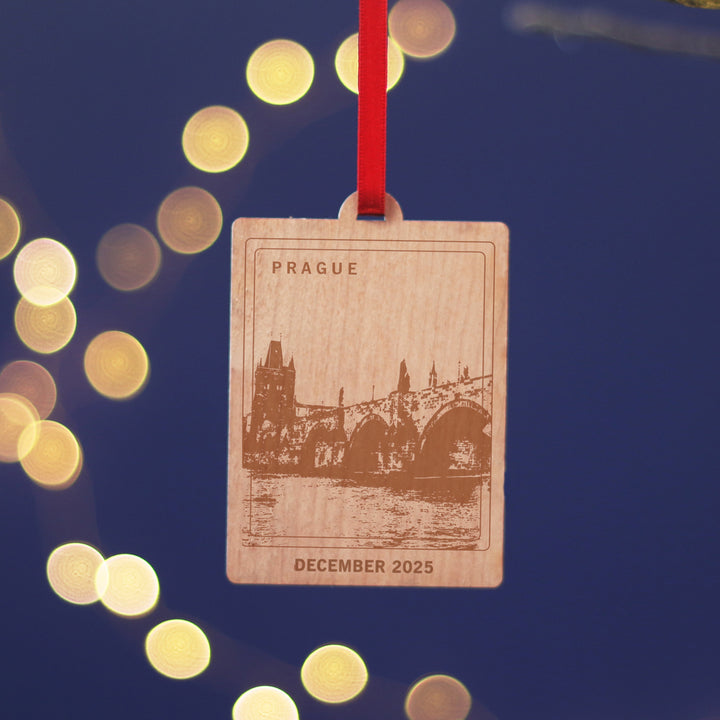 Prague travel memory anniversary gift, commemorate your Christmas memories, with this sustainable wood cherry veneered ornament with the Charles Bridge Hanging against a blue background. Personalise the location and text for a perfect letterbox friendly gift for travellers.