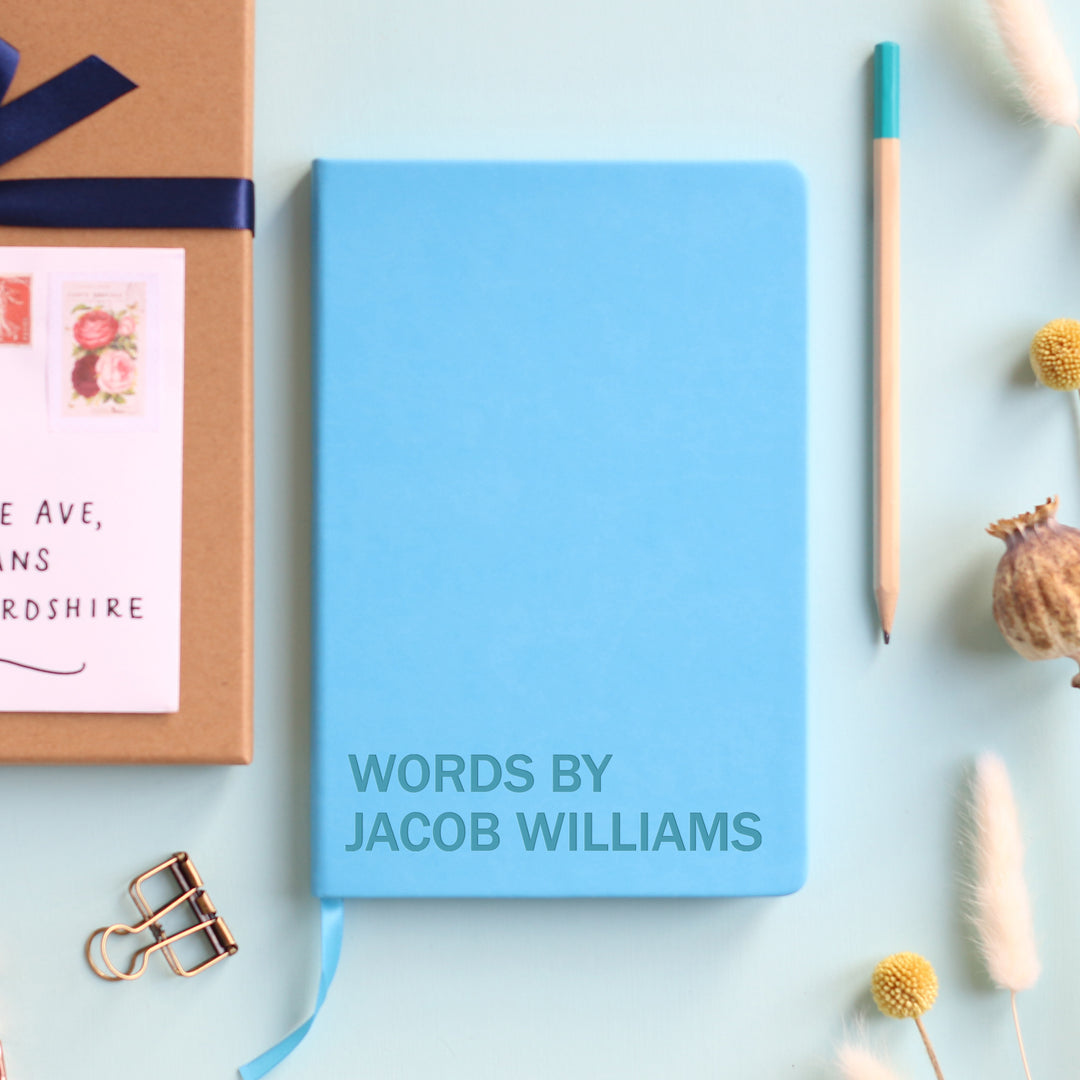 A top down image of a blue vegan leather resting on a pale aqua table. The front of the notebook is  engraved with the phrase Words By and personalised with the name Jacob Williams. Surrounding the book are various stationary items and dried flowers.