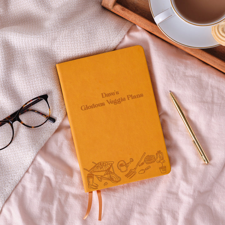 A top down image of a mustard yellow vegan leather notebook. The front of the notebook features an engraving of gardening tools along the bottom edge.  The personalised engraving reads “Dave’s Glorious Veggie Plans”. Surrounding the book is a tray with a cup of tea and a biscuit, a pair of glasses and a gold pen.