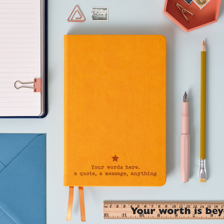 A top down image of a Mustard Yellow hardcover vegan leather notebook resting on a pale aqua table. The bottom of the book is engraved with a personalised message, Your words here. a quote, a message, anything and a solid star symbol  is engraved above the text. Surrounding the book are various stationary.
