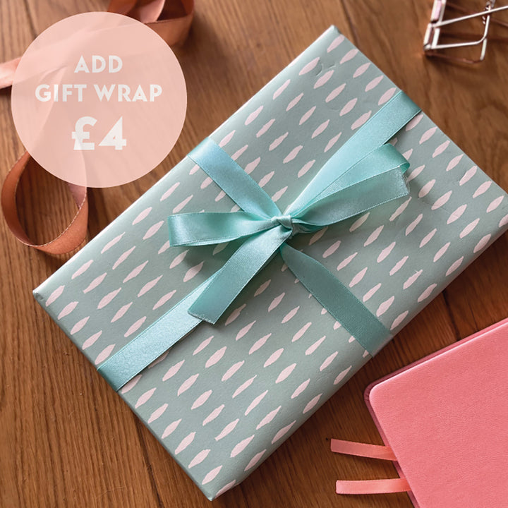 An A5 notebook is wrapped in a pale aqua gift wrap with white pattern. Finished with a matching aqua satin ribbon. Placed next to a pink notebook. The text ADD GIFT WRAP £4 is written in a circle on the top left corner of the image.