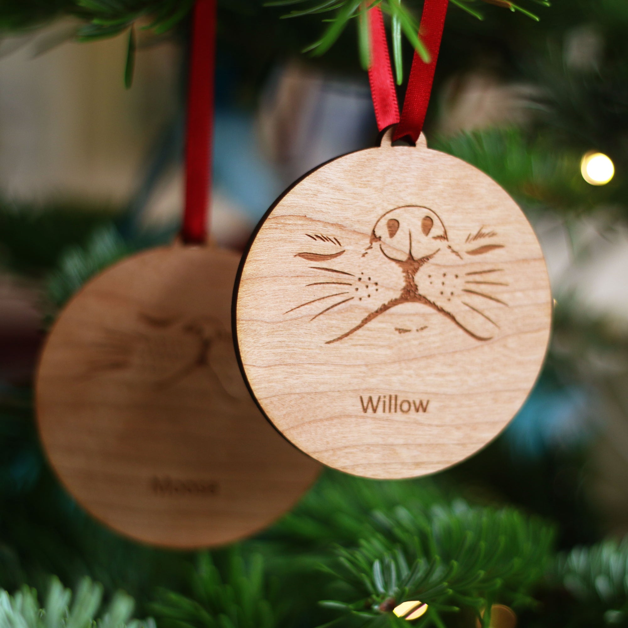A pair of wooden pet Christmas tree baubles hanging together on a tree. One featuring a dog nose, the other featuring a cat nose. Both personalised with the pet's name.