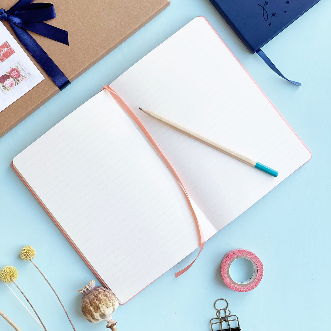 Personalised journal notebook lying flat open on pale aqua table to show lined pages and a matching colour ribbon place marker. A pencil is resting on the top of the notebook. Washi tape, clips and other stationery items are scattered around it. 