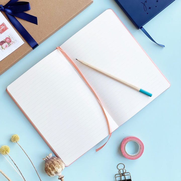 Personalised journal notebook lying flat open on pale aqua table to show lined pages and a matching colour ribbon place marker. A pencil is resting on the top of the notebook. Washi tape, clips and other stationery items are scattered around it. 