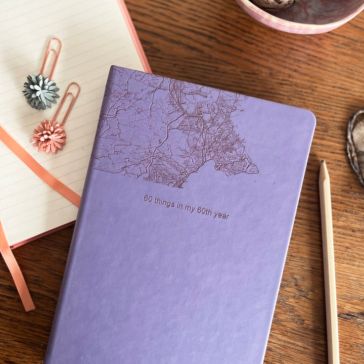 A top down image of a lilac vegan leather notebook resting on a wooden table. The top third of the notebook is engraved with a map and personalised with the text “60 things in my 60th year”. Surrounding the book are various stationery items and dried flowers.