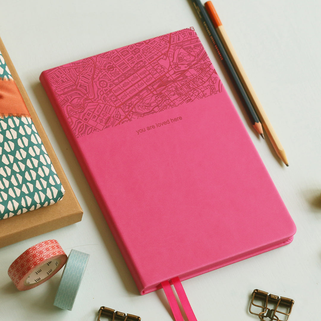 A top down image of a hot pink vegan leather notebook resting on a pale aqua table. The top third of the notebook is engraved with a map and personalised with the text “you are loved here”. Surrounding the book are various stationary items.