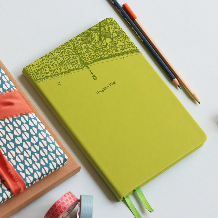 A top down image of a lime green vegan leather notebook resting on a pale aqua table. The top third of the notebook is engraved with a map and personalised with the text “Brighton Pier”. Surrounding the book are various stationary items.
