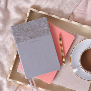 A top down image of a grey vegan leather notebook. The top third of the notebook is engraved with a map and personalised with the text “my happy place”.  It is stacked above a pink notebook and is resting on a cream wooden tray with a cup of tea, and a gold pen beside it. The tray is resting on a pink bedspread.