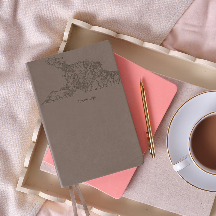 A Top down view of a grey notebook resting on a pink notebook on a tray with a cup of tea. The notebook has a map engraved on the top third with the text 'happy days' engraved below. The tray is sitting on a soft pink bedspread.