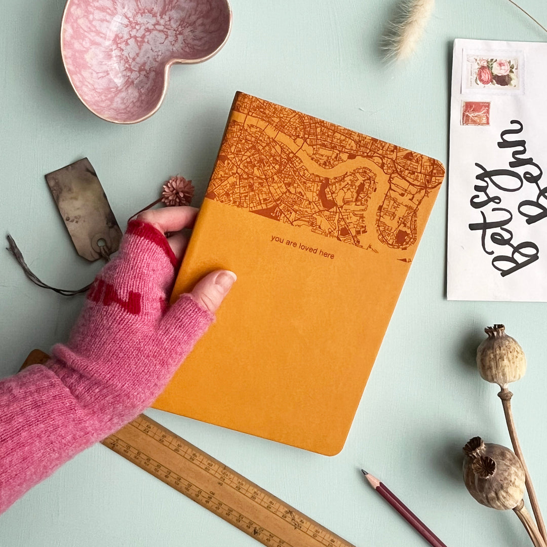 A close up image of a mustard yellow vegan leather notebook being held by a hand wearing a pink fingerless glove. The background is a pale aqua table. The top third of the notebook is engraved with a map and personalised with the text “you are loved here”. Surrounding the book are various stationary items and dried flowers.