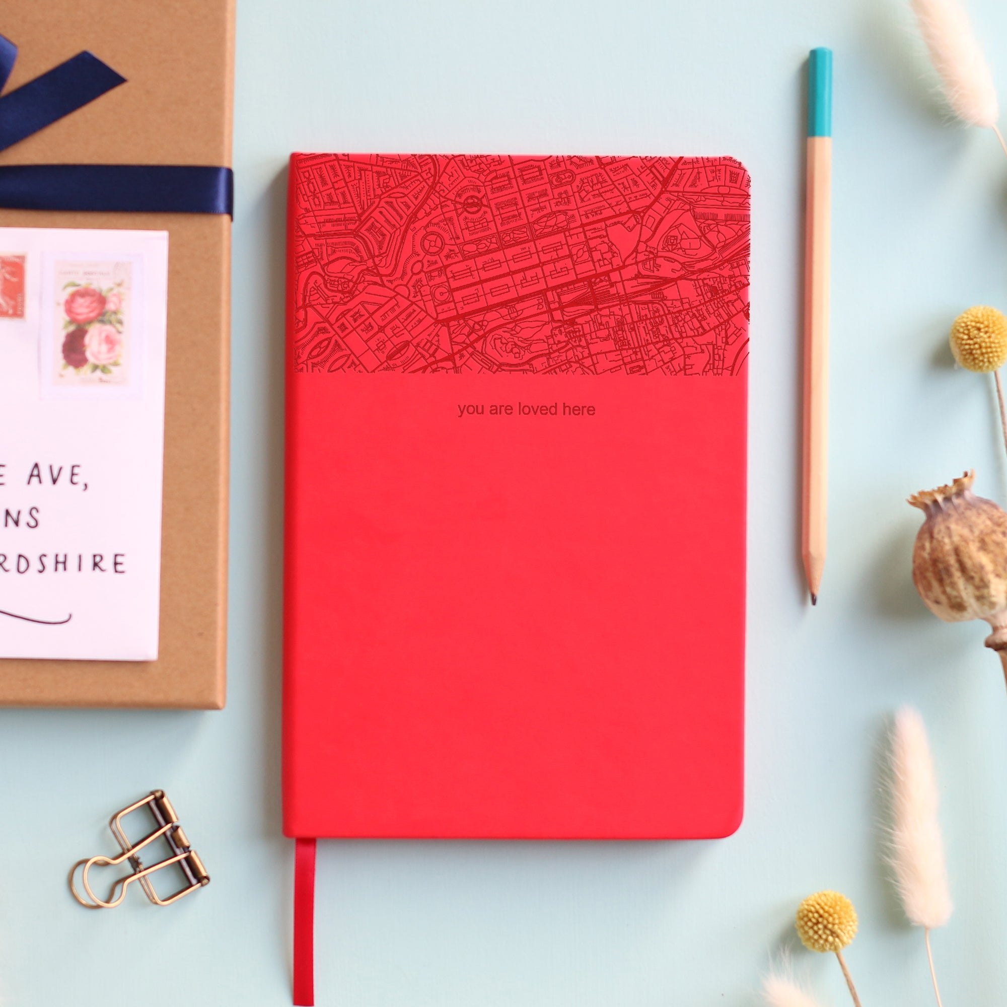 A top down image of a red vegan leather notebook resting on a pale aqua table. The top third of the notebook is engraved with a map and personalised with the text “you are loved here”. Surrounding the book are various stationary items and dried flowers.